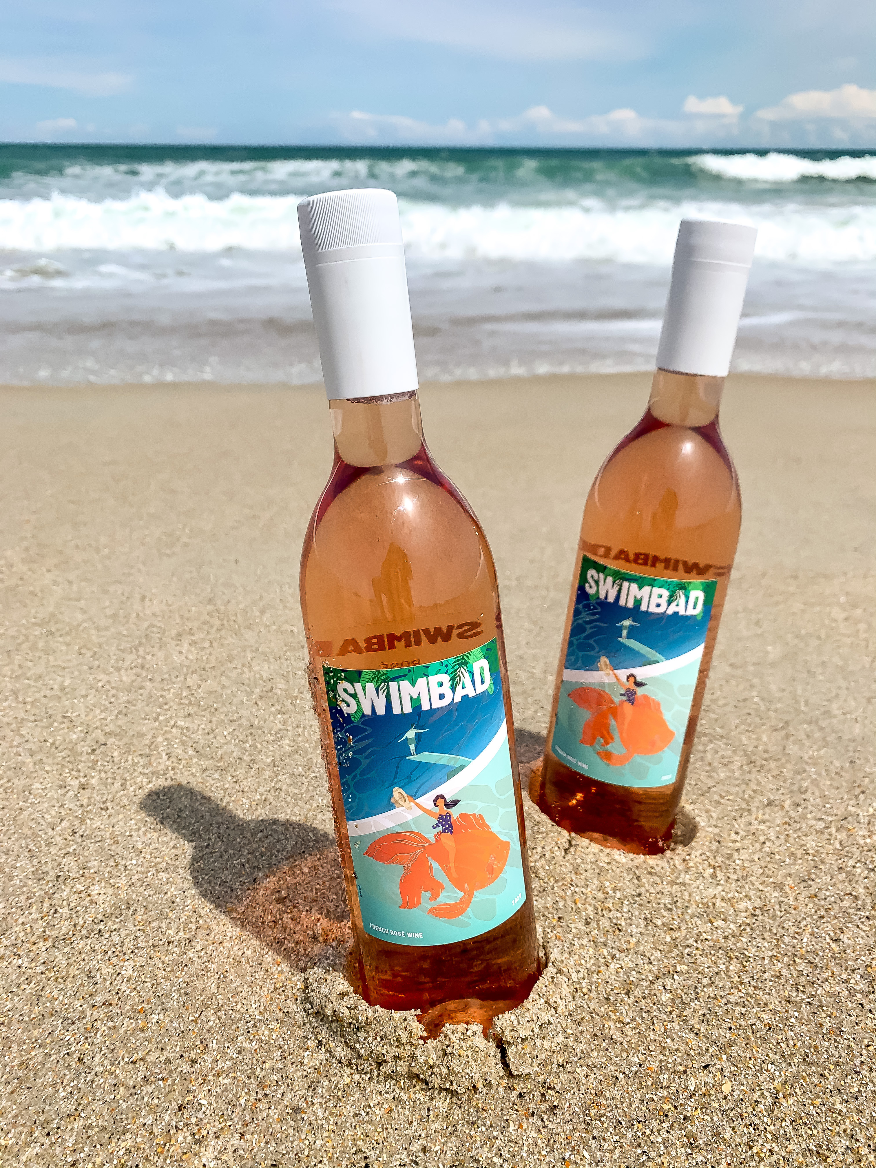 Two bottles of Swimbad rosé standing in beach sand