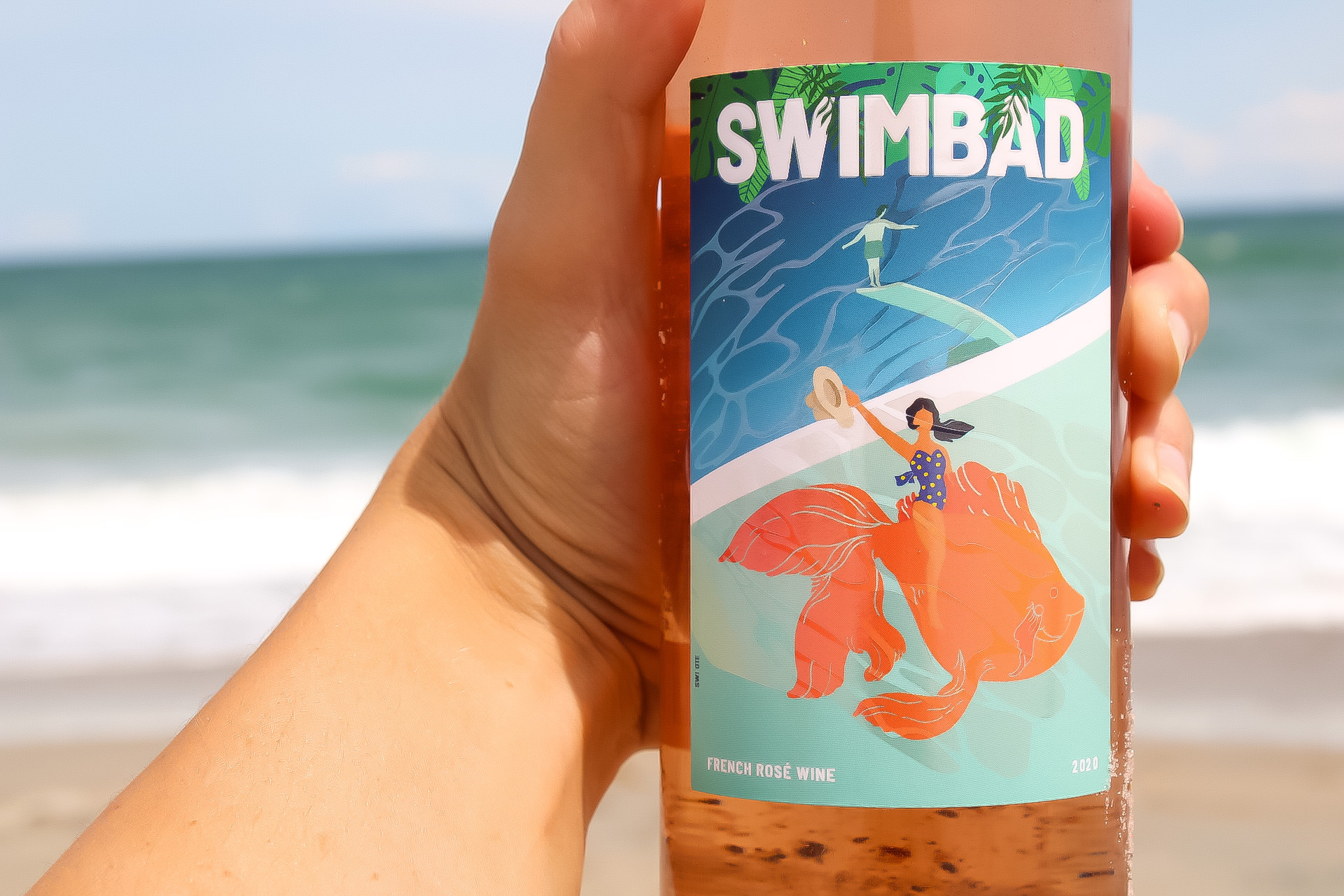 A hand holding a bottle of Swimbad rosé in front of the ocean